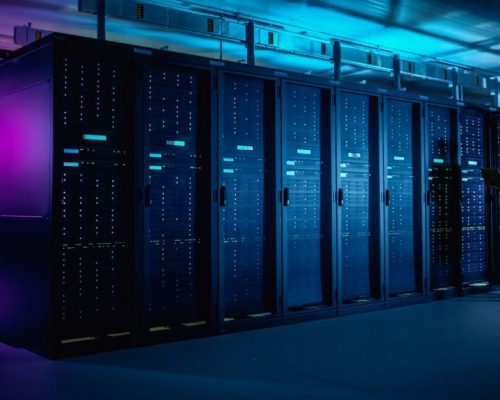 In Data Center: IT Technician Running Maintenance Programme on Laptop, Controls Operational Server Rack Optimal Functioning. Modern High-Tech Telecommunications Operational Data Center in Neon Lights.