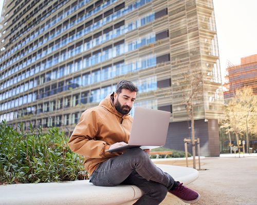 Business man typing on laptop computer outdoors. Casual dressed man working with laptop sitting on a bench. Concept of telecommuting. Remote work.